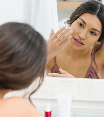 Woman applying facial moisturizer to her face on front of a bathroom mirror.
