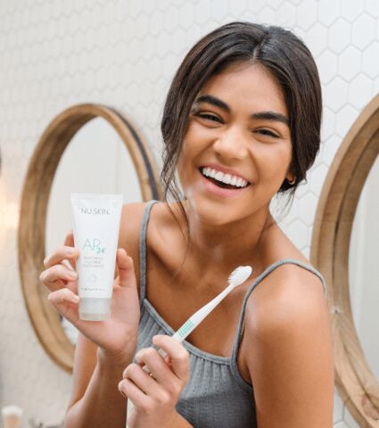 Young woman smiling while holding toothbrush and AP24 whitening fluoride toothpaste in bathroom.