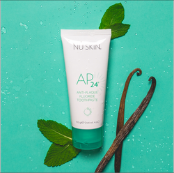 A bottle of the AP 24 anti-plaque fluoride toothpaste laying on a green background with mint and vanilla leaves around it. 