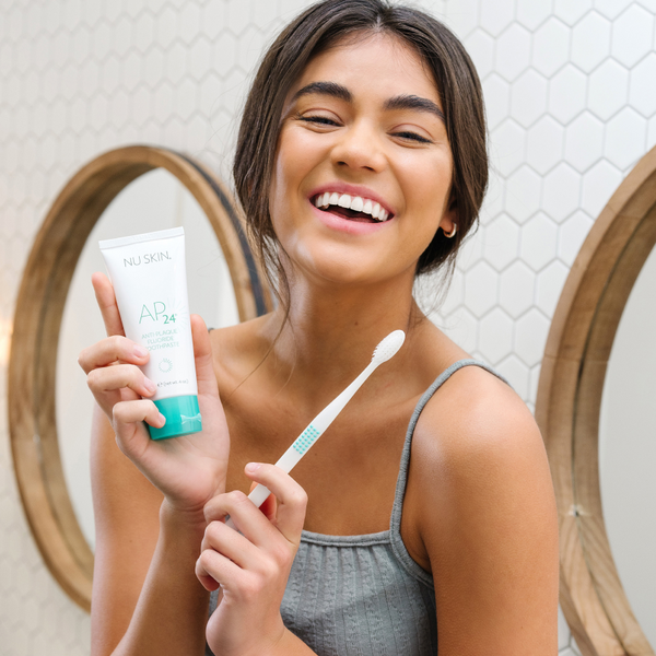 A woman smiling while holding the AP 24 Anti-plaque fluoride toothpaste in one hand and a toothbrush on the other hand.