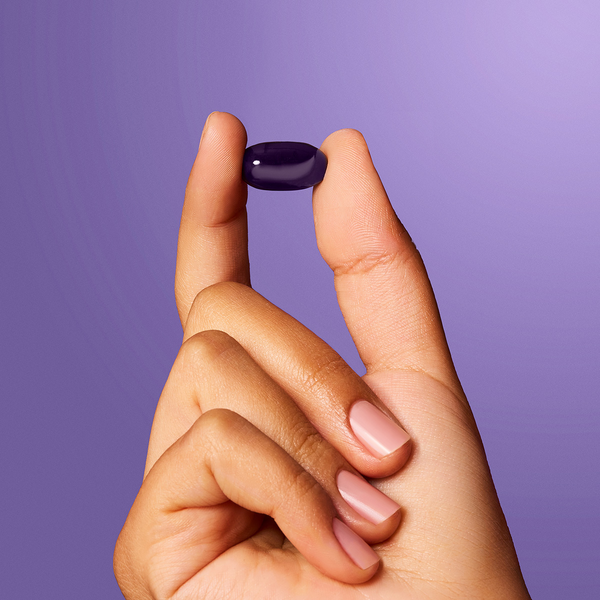 Two fingers holding up an ageLOC Meta supplement pill in front of a purple backdrop.